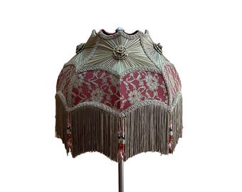 Puede incluir: Una pantalla de lámpara ornamentada con un patrón de encaje rojo y ribetes dorados. La pantalla presenta un borde festoneado, flecos y detalles florales decorativos. Los flecos están adornados con pequeñas cuentas de colores.