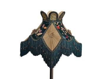 May include: An ornate lampshade with a dark teal fringe and floral accents. The shade features a diamond-shaped center with a rosette design and is trimmed with a gold-colored border. The top of the shade has a scalloped edge.