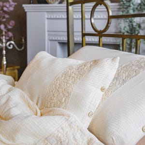 May include: Cream-colored bedding set featuring a textured duvet and pillows. The pillows have a decorative lace trim and button details. The bed frame is gold-toned, adding a touch of elegance to the bedroom decor.