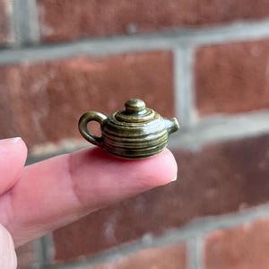 May include: A miniature ceramic teapot with a glossy olive green glaze and horizontal ribbed detailing. The small vessel features a rounded body, a short spout, a curved handle, and a matching lid with a small knob on top, resting on a fingertip.