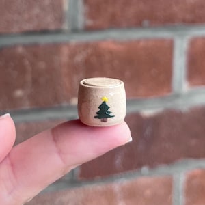 May include: A miniature cylindrical ceramic container with a smooth beige finish. The exterior features a hand-painted dark green pine tree graphic topped with a small yellow star. The object has a flat, circular lid that sits flush on top.