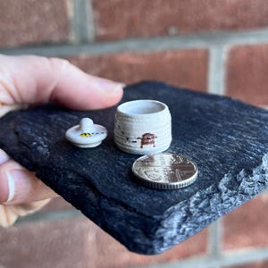 May include: A miniature ceramic jar with a lid, decorated with a bee design, sits on a dark slate surface. The jar is cream-colored with brown and black accents. A coin is next to the jar for size comparison. The jar is likely handmade.