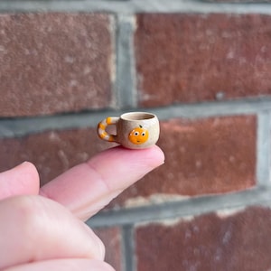 Może przedstawiać: Miniaturowy ceramiczny kubek z motywem dyni. Kubek jest beżowy z pomarańczowo-żółtym uchwytem. Twarz dyni jest pomarańczowa z uśmiechniętą miną. Kubek jest trzymany na palcu.