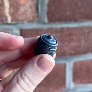 May include: A small, dark blue bottle stopper with a rounded top and a cylindrical body. The stopper has a ridged texture and a small, circular knob on top. The object is held between fingers.