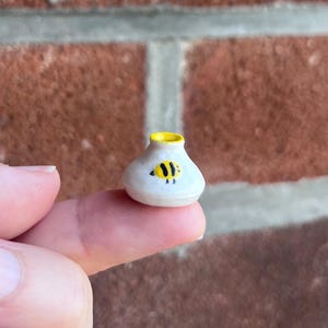 Puede incluir: Un jarrón de cerámica en miniatura con un acabado blanco brillante, que presenta un pequeño motivo de abeja amarillo y negro pintado a mano en la parte frontal y un borde amarillo brillante alrededor de la abertura. El objeto es redondeado con un cuello estrecho y una base ancha.