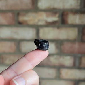Pode incluir: Uma caneca de cerâmica preta em miniatura com uma pequena alça e detalhes de pontos brancos. A pequena caneca é segurada em um dedo, mostrando seu tamanho pequeno. O fundo é uma parede de tijolos.