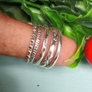 Può includere: Una collezione di anelli d'argento con vari design, tra cui fasce strutturate e lisce. Gli anelli sono impilati su un dito, mostrando i loro diversi stili. L'immagine è ambientata su uno sfondo di fogliame verde e un oggetto rosso.