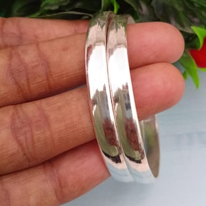 Puede incluir: Dos pulseras de plata pulida. Las pulseras son lisas y reflectantes, con una superficie brillante y reluciente. Las joyas se exhiben sobre un fondo de hojas verdes y una superficie azul claro.