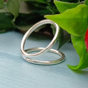 Könnte beinhalten: Ein silberner Ring mit einem einzigartigen Doppelband-Design. Der Ring hat eine glatte, polierte Oberfläche und eine moderne, minimalistische Ästhetik. Der Ring ist vor einem hellblauen Hintergrund mit grünem Laub platziert.