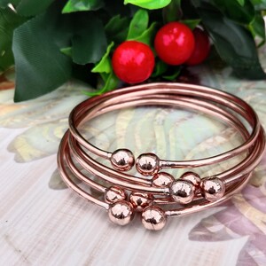 Könnte beinhalten: Mehrere roségoldene Armreifen mit kugelförmigen Details. Die Armbänder sind auf einer hellen Oberfläche gestapelt, mit roten Beeren und grünen Blättern im Hintergrund. Schlichtes, elegantes Design.