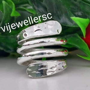 May include: A silver-colored, hammered-finish ring in a spiral design. The ring wraps around the finger multiple times, with a textured surface. The background features green foliage and a red flower, with the text "urvijewellersc".