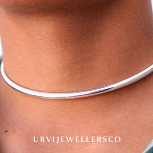 以下が含まれることがあります： 光沢のあるシルバーカラーのチョーカーネックレス。 ネックレスはシンプルで滑らかで丸みを帯びたバンドです。 ネックレスは首の周りに着用されています。 画像の下部には「URVIJEWELLERSCO」というテキストが表示されています。