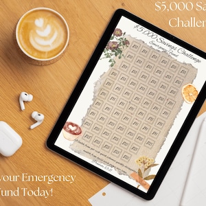 May include: A digital tablet displays a $5,000 Savings Challenge chart with boxes labeled $50 and $25. The text "Start your Emergency Fund Today!" is at the bottom. A cup of coffee, wireless earbuds, and a case are on a wooden surface.