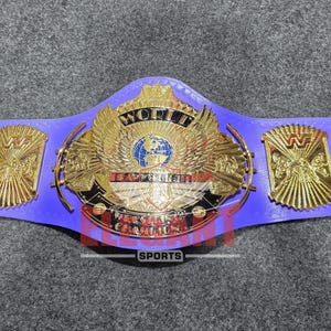 Puede incluir: Un cinturón de campeonato de lucha libre con una correa de cuero morado brillante y cinco placas de metal dorado. La placa central grande muestra un globo terráqueo, alas y el texto WORLD HEAVYWEIGHT CHAMPION. Cuatro placas laterales llevan el logo WWF.