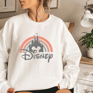 May include: White sweatshirt featuring a colorful rainbow design with a castle silhouette and the word "Disney" in a script font. The design incorporates pastel colors and a Mickey Mouse head. A casual, comfortable top.