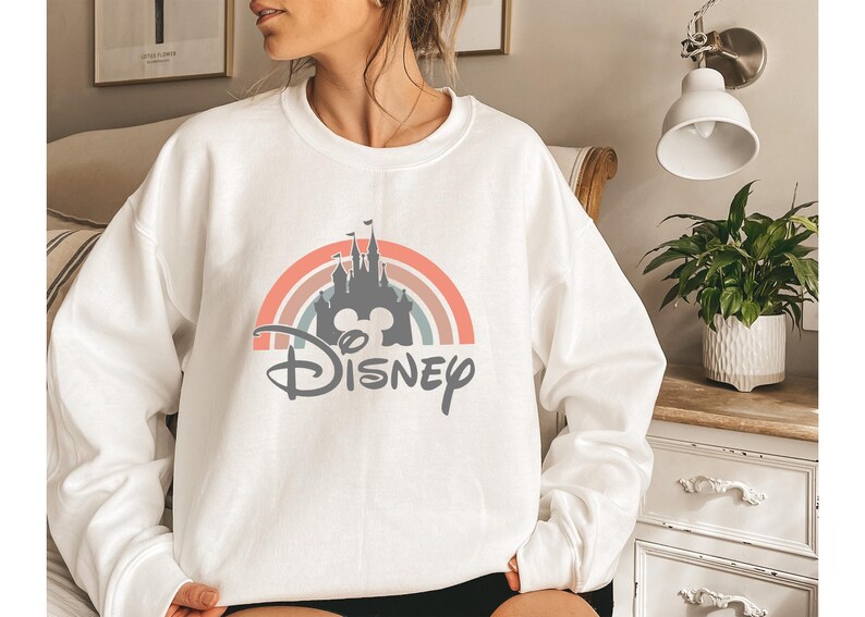 May include: White sweatshirt featuring a colorful rainbow design with a castle silhouette and the word "Disney" in a script font. The design incorporates pastel colors and a Mickey Mouse head. A casual, comfortable top.