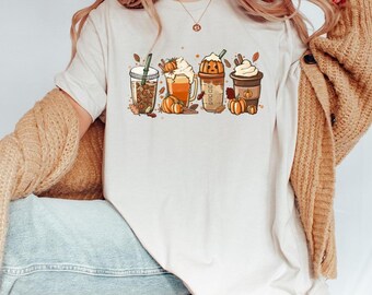 May include: Cream-colored t-shirt featuring a fall-themed graphic of four different pumpkin spice lattes with pumpkins and autumn leaves. The shirt is worn by a person wearing a tan cardigan and blue jeans.