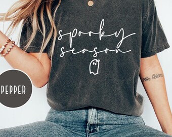 May include: Dark grey t-shirt with the words "spooky season" in white script and a small ghost graphic. The shirt is paired with blue jeans. The word "pepper" is in a circle on the left side of the image.