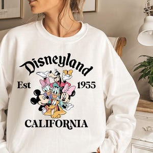 Puede incluir: Sudadera blanca con el logotipo de Disneyland y los personajes de dibujos animados Mickey Mouse, Minnie Mouse, Donald Duck, Daisy Duck, Goofy y Pluto. El texto "Est 1955 CALIFORNIA" tambi&eacute;n est&aacute; impreso.