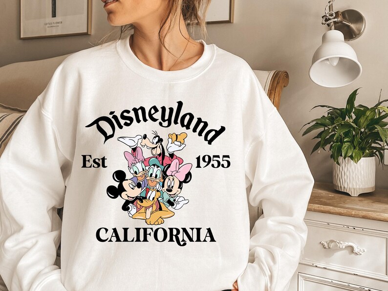 Puede incluir: Sudadera blanca con el logotipo de Disneyland y los personajes de dibujos animados Mickey Mouse, Minnie Mouse, Donald Duck, Daisy Duck, Goofy y Pluto. El texto "Est 1955 CALIFORNIA" tambi&eacute;n est&aacute; impreso.