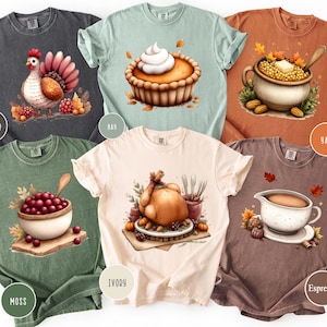 May include: A collection of six t-shirts in various colors, each featuring a watercolor illustration of Thanksgiving-themed food items. Designs include a turkey, pie, and cranberry sauce. The shirts are labeled with color names like "Pepper" and "Ivory".