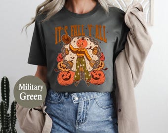 May include: Military green t-shirt with the text "IT'S FALL Y'ALL" in orange. The graphic features a pumpkin-headed scarecrow holding a hat, surrounded by pumpkins and chains. The shirt is paired with denim shorts.