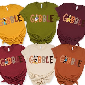 May include: A collection of six short-sleeved t-shirts in various colors, each featuring a different Disney character and the word "GOBBLE" in a playful font. The shirts are in shades of yellow, green, burgundy, tan, and orange.