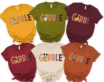 May include: A collection of six short-sleeved t-shirts in various colors, each featuring a different Disney character and the word "GOBBLE" in a playful font. The shirts are in shades of yellow, green, burgundy, tan, and orange.