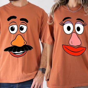 May include: Two rust-colored t-shirts, each featuring a cartoon face design. One shirt has a mustache and a smiling mouth, while the other has eyelashes and a red lip. The shirts are short-sleeved and appear to be made of a soft material.