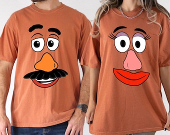May include: Two rust-colored t-shirts, each featuring a cartoon face design. One shirt has a mustache and a smiling mouth, while the other has eyelashes and a red lip. The shirts are short-sleeved and appear to be made of a soft material.