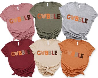 May include: A collection of six t-shirts in various colors, each featuring the word "GOBBLE" in a playful font. The letter "O" is replaced with a Mickey Mouse head silhouette. The shirts are in shades of pink, green, gray, burgundy, beige, and orange.