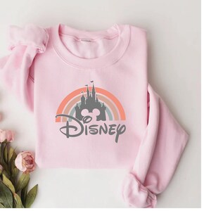 May include: A light pink sweatshirt featuring a colorful rainbow design with a castle silhouette and the word "Disney" in a script font. The sweatshirt is folded neatly, with a bouquet of pink flowers in the corner.