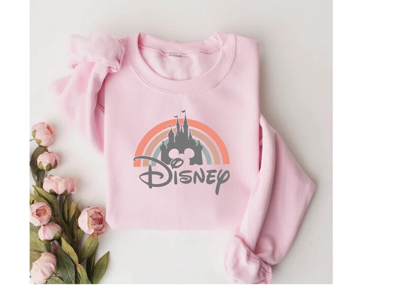 May include: A light pink sweatshirt featuring a colorful rainbow design with a castle silhouette and the word "Disney" in a script font. The sweatshirt is folded neatly, with a bouquet of pink flowers in the corner.