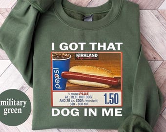 May include: Military green sweatshirt with the text "I GOT THAT DOG IN ME." The graphic features a hot dog, soda, and a price tag. The sweatshirt is a solid color with long sleeves.