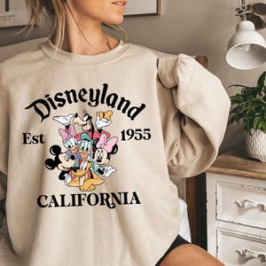 Puede incluir: Sudadera beige de Disneyland con Mickey Mouse, Minnie Mouse, Donald Duck, Daisy Duck, Goofy y Pluto. La sudadera tiene las palabras "Disneyland Est 1955 California" en escritura negra.