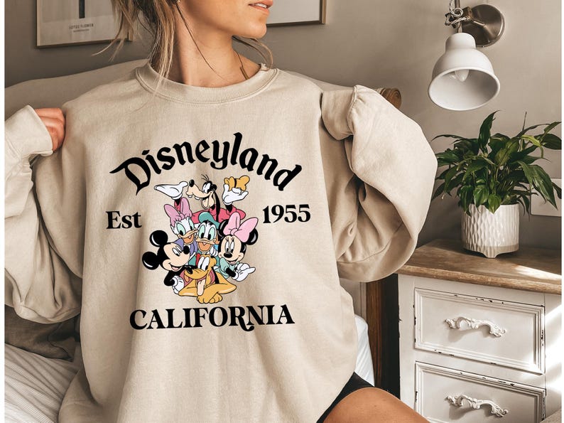 Puede incluir: Sudadera beige de Disneyland con Mickey Mouse, Minnie Mouse, Donald Duck, Daisy Duck, Goofy y Pluto. La sudadera tiene las palabras "Disneyland Est 1955 California" en escritura negra.