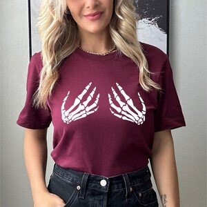 May include: A maroon t-shirt featuring a white graphic of two skeleton hands. The short-sleeved shirt is made of a soft material and is suitable for casual wear. The design is centered on the chest, creating a unique and eye-catching look.
