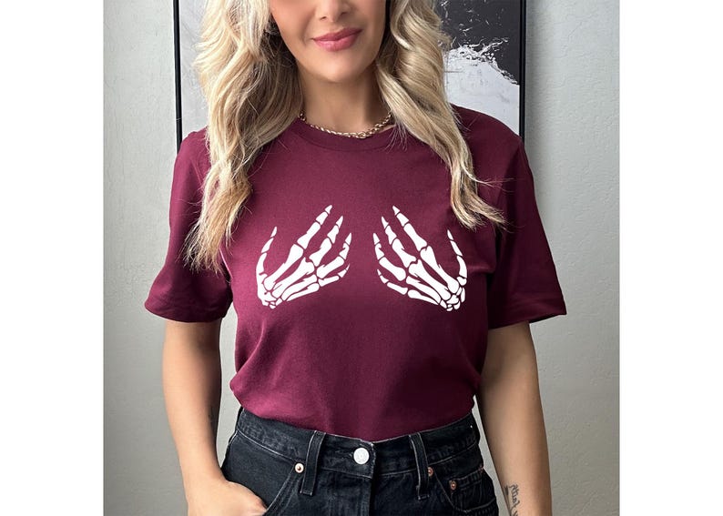May include: A maroon t-shirt featuring a white graphic of two skeleton hands. The short-sleeved shirt is made of a soft material and is suitable for casual wear. The design is centered on the chest, creating a unique and eye-catching look.