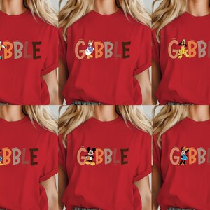 May include: Red t-shirts with the word "GOBBLE" in various colors and cartoon characters. The characters include Goofy, Donald Duck, Pluto, Daisy Duck, Mickey Mouse, and Minnie Mouse. The shirts are short-sleeved.