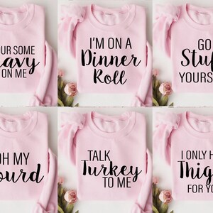 May include: Six light pink sweatshirts are displayed, each with a different black text slogan. The phrases read: "Pour Some Gravy On Me," "I'm On A Dinner Roll," "Go Stuff Yourself," "Oh My Gourd," "Talk Turkey To Me," and "I Only Have Thighs For You."