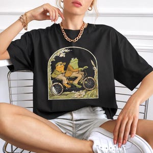 Puede incluir: Camiseta negra con una ilustraci&oacute;n caprichosa de dos ranas en bicicleta. Las ranas visten ropa de &eacute;poca, sobre un fondo de follaje dentro de un marco arqueado. Una camiseta gr&aacute;fica divertida y &uacute;nica.