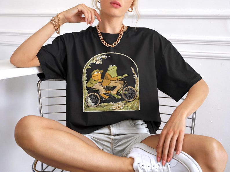 Puede incluir: Camiseta negra con una ilustraci&oacute;n caprichosa de dos ranas en bicicleta. Las ranas visten ropa de &eacute;poca, sobre un fondo de follaje dentro de un marco arqueado. Una camiseta gr&aacute;fica divertida y &uacute;nica.
