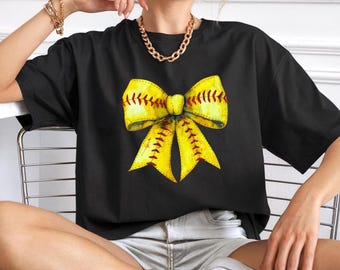 May include: Black t-shirt with a large yellow bow graphic, detailed with red stitching, resembling a softball. The shirt has a relaxed fit, suitable for casual wear. The design is eye-catching and sporty.