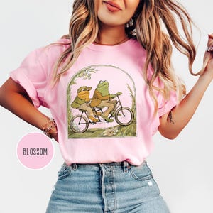 Puede incluir: Camiseta rosa con una ilustraci&oacute;n caprichosa de dos ranas en bicicleta t&aacute;ndem. Las ranas visten ropa vintage, enmarcada en un arco con detalles florales. La palabra "Blossom" est&aacute; en un c&iacute;rculo rosa.