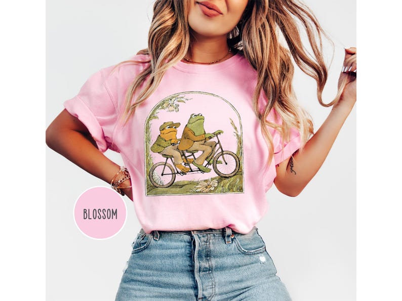 Puede incluir: Camiseta rosa con una ilustraci&oacute;n caprichosa de dos ranas en bicicleta t&aacute;ndem. Las ranas visten ropa vintage, enmarcada en un arco con detalles florales. La palabra "Blossom" est&aacute; en un c&iacute;rculo rosa.