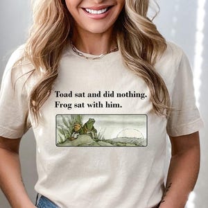 May include: Beige t-shirt featuring the text "Toad sat and did nothing. Frog sat with him." and an illustration of two frogs. The illustration shows a frog and toad sitting together. The shirt is a neutral color.