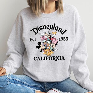 Puede incluir: Una sudadera gris claro con un gr&aacute;fico de Disneyland. El gr&aacute;fico incluye a Mickey Mouse, Minnie Mouse, Donald Duck, Daisy Duck, Goofy y Pluto. El texto dice "Disneyland Est 1955 California".