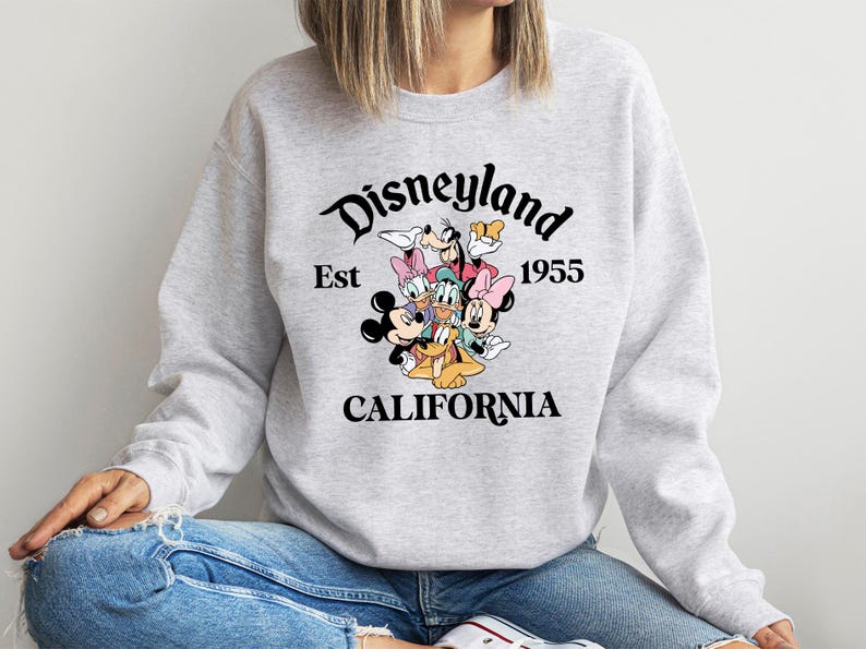Puede incluir: Una sudadera gris claro con un gr&aacute;fico de Disneyland. El gr&aacute;fico incluye a Mickey Mouse, Minnie Mouse, Donald Duck, Daisy Duck, Goofy y Pluto. El texto dice "Disneyland Est 1955 California".