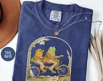 May include: True navy blue t-shirt featuring a whimsical illustration of two frogs riding a bicycle. The frogs are dressed in vintage clothing, and the design is framed by an archway of flowers and foliage. The shirt has a Comfort Colors label.