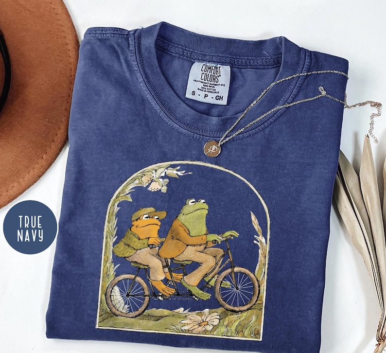 Puede incluir: Camiseta azul marino con una ilustraci&oacute;n caprichosa de dos ranas en bicicleta. Las ranas visten ropa vintage, y el dise&ntilde;o est&aacute; enmarcado por un arco de flores y follaje. La camiseta tiene una etiqueta Comfort Colors.
